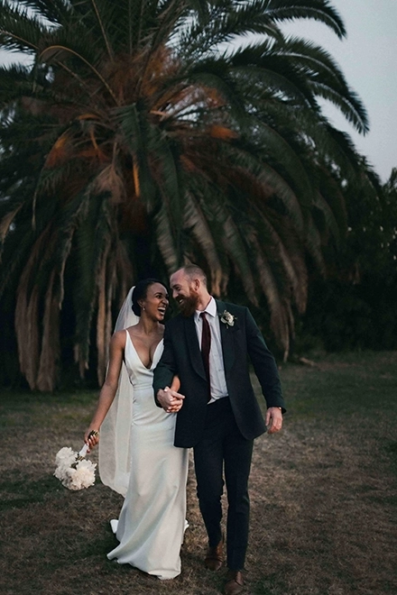 Photographie d'un couple mixte de jeunes mariés, se tenant la main et riant en se regardant.